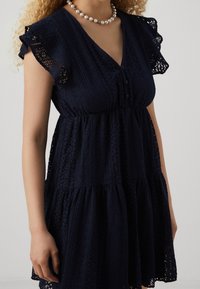 Granatowa sukienka z teksturowanym koronkowym wzorem, krótki rękaw z falbanką oraz zapinany na guziki dekolt. Lekko rozkloszowany dół i marszczona talia.
