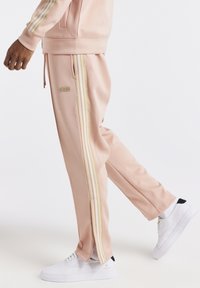 Světle růžové tepláky s bílými a zlatými bočními pruhy, vyrobené z hladké látky. Mají šňůrku v pase a zipy na kotnících.