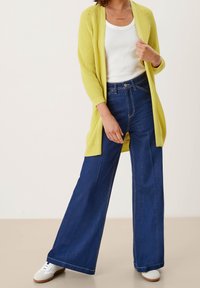 Dlouhý žlutý pletený kardigan přes bílý tílko, spárovaný s volnými tmavě modrými džíny a bílými teniskami s béžovými podrážkami. Detaily neformálního outfitu.