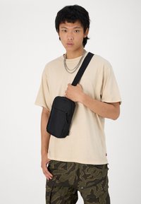 Zwarte stoffen schoudertas met een hoofdcompartiment met rits, voorvak en verstelbare schouderband. Draagt over een beige t-shirt en camouflagebroek.