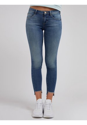 Jean slim bleu en denim avec ourlets effilochés aux chevilles porté avec des baskets blanches sur une personne debout sur un fond uni.