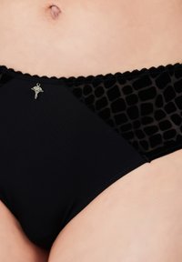 Schwarze Panties mit einem durchsichtigen Mesh-Einsatz und einem strukturierten Tiermuster, verziert mit einem kleinen dekorativen Anhänger und zarter, gewellter Borte.