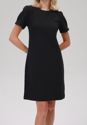 Femme portant une robe noire texturée à manches courtes, longueur genoux, debout, mains dans le dos, sur un fond clair uni.