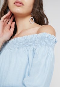 Jasnoniebieska bluzka z odkrytymi ramionami, z marszczonym dekoltem i falbankami na brzegach, zestawiona z dużymi przezroczystymi kolczykami-kołami.