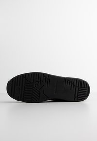 Black rubber shoe sole with textured pattern, featuring horizontal and vertical grooves for traction and a circular logo near the heel.