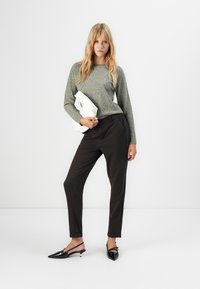 Woman standing with loose gray sweater, black tailored pants, black slingback heels, and holding a white clutch against a plain background.
