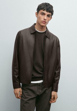 Junger Mann, der eine dunkelbraune Lederjacke über einem braunen Pullover und grauen Jeans trägt, steht vor einem schlichten hellgrauen Hintergrund.