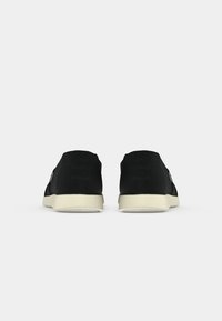 Paire de chaussures noires type slip-on avec semelles blanches, vues de dos sur un fond clair uni.