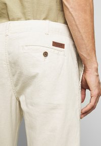 Pantalones de algodón beige con un bolsillo trasero que presenta una parche de cuero marrón etiquetado como "INDICODE". La textura es suave con detalles de costura visibles.