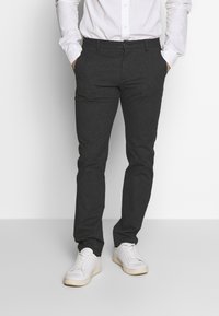 Muž oblečený v tmavě šedých kalhotách, bílé košili a bílých teniskách stojí s rukama v kapsách na jednobarevném pozadí.