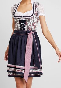 Femme portant une robe traditionnelle bavaroise dirndl avec des manches en dentelle, un corsage à motifs floraux, une jupe bleu marine à pois blancs et un nœud à carreaux roses à la taille.