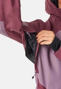 Jas in tinten van kastanjebruin en lavendel met een ritszak, flexibel materiaal en aanpassingsmogelijkheden op de manchetten en capuchon.