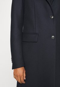 Marineblauwe wollen jas met een gestructureerde afwerking, enkelvoudige knopen, en twee knopen. Beschikt over een zijzak en gegoten mouwen.