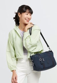 Light green cropped jacket with elastic cuffs, worn over a gray top; carrying a navy blue bag with zippers and a shoulder strap.