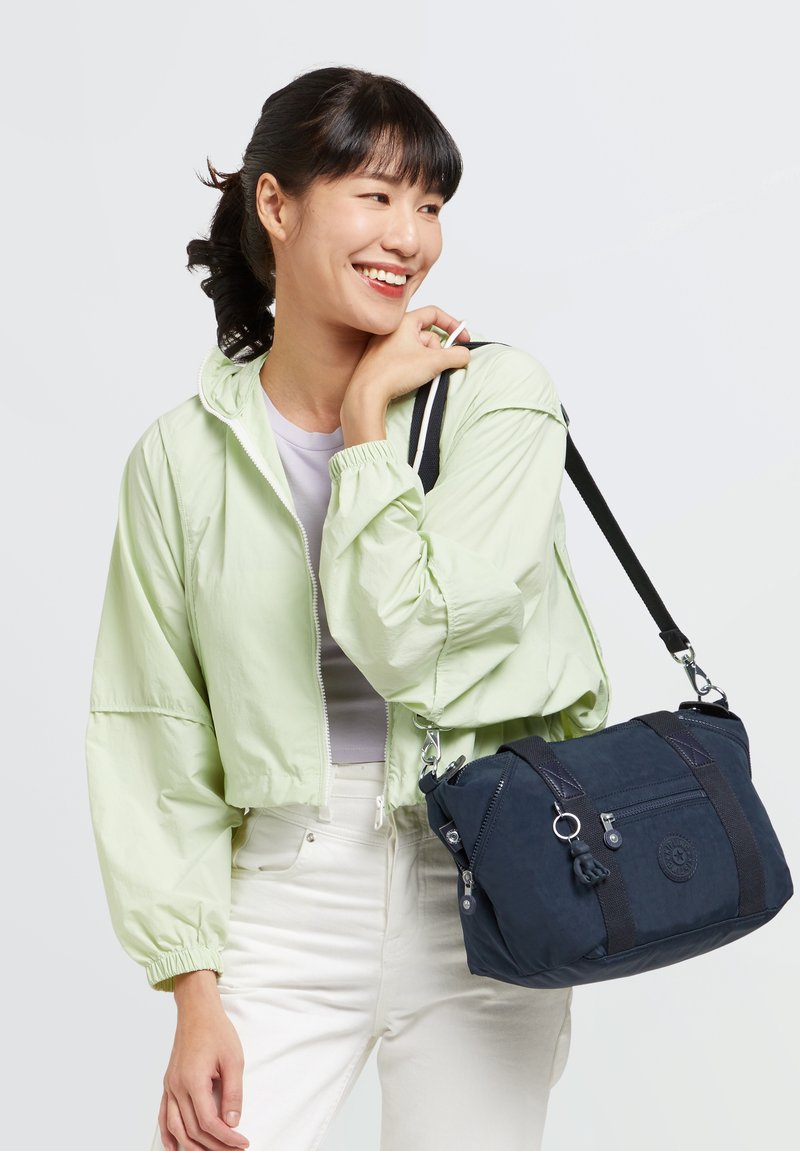 Light green cropped jacket with elastic cuffs, worn over a gray top; carrying a navy blue bag with zippers and a shoulder strap.