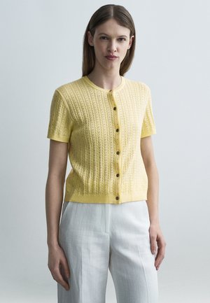 Femeie cu păr drept și castaniu, purtând un top tricotat galben cu mâneci scurte și nasturi, și pantaloni albi cu talie înaltă, pe un fundal simplu.