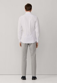 Camicia bianca a quadri con colletto standard, maniche lunghe e orlo arrotondato, abbinata a pantaloni grigi chiari e scarpe scure.