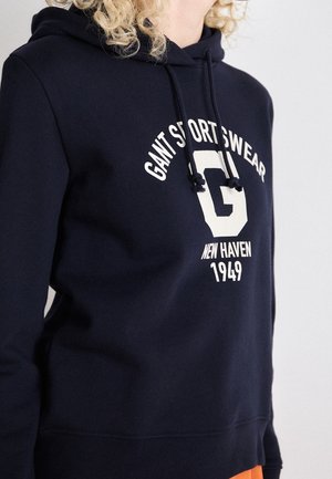 Persoon draagt een marineblauwe GANT Sportswear hoodie met een wit logo en tekst "New Haven 1949", met deels zichtbaar krullend blond haar.