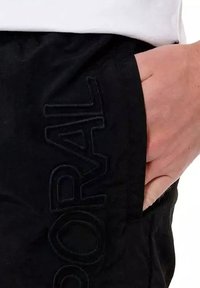 Shorts noirs avec un logo brodé en relief sur la poche latérale, une texture lisse et une coupe décontractée. La main du modèle repose dans la poche.
