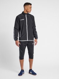 Schwarze Sportjacke mit weißem Reißverschluss und Logo, mit Chevron-Muster-Akzenten auf den Ärmeln. Passende schwarze Cropped-Hosen. Blaue Turnschuhe.