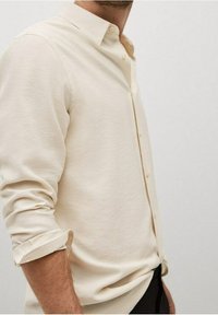 Homme portant une chemise à manches longues crème, manches retroussées, sur un fond blanc uni.