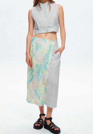 Haut gris texturé coupé avec un col haut, associé à une jupe à motifs pastel avec un détail de bouton sur le côté. Sandales noires à lanières.