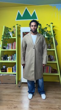 Bēšs vilnas mētelis ar vienu pogu aizdari, atloku apkakli un divām sānu kabatām, valkāts kopā ar zilajām džinsa biksēm un baltām sporta kurpēm.