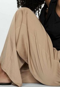 Pantalon beige à jambes larges en tissu doux, avec une coupe fluide et des plis subtils. Associé à un haut noir et des sandales.