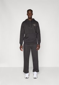 Mørkegrå hoodie med frontlomme og hvidt logo, parret med matchende bukser. Har snore og elastiske manchetter, båret med hvide sneakers.