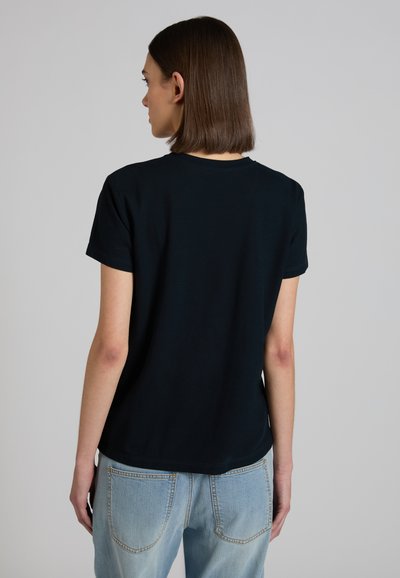 Vrouw met stijlbruin haar, een eenvoudige zwarte korte mouw t-shirt en lichtblauwe spijkerbroek, van achteren gezien tegen een effen grijze achtergrond.