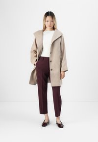 Femme se tenant debout, portant un manteau beige, un haut blanc, un pantalon bordeaux et des mocassins bordeaux foncé sur fond blanc.