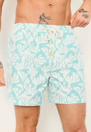 Hellblaue Badeshorts mit weißen abstrakten Blattmustern und einem weißen Kordelzug, getragen von einer Person mit Tätowierungen an Hand und Bauch.