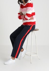 Pull rayé rouge et rose avec un ourlet côtelé, associé à un pantalon bleu marine avec une bande latérale rouge, et des baskets blanches sur un tabouret.