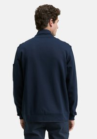 Homme avec des cheveux bouclés portant une veste à manches longues bleu marine et un pantalon assorti, se tenant de dos contre un fond uni.