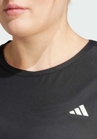 Mustat urheilu-paita, jossa on teksturoitu kangas, pyöreä kaula-aukko ja valkoinen vinosti asetettu logo vasemmassa rintamuksessa.