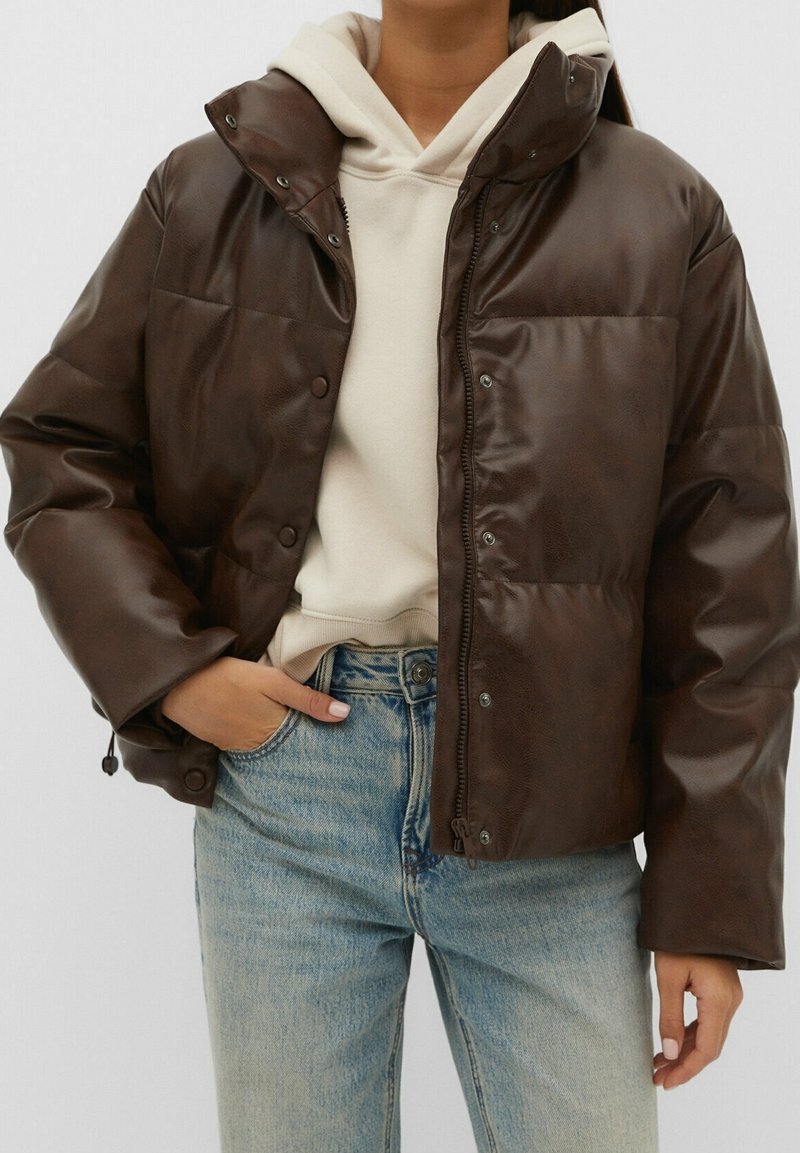 Bruine nep leren pufferjack met een ritssluiting aan de voorkant, drukknoopsluiting en een semi-gekort model, gedragen over een beige hoodie en lichtblauwe spijkerbroeken.