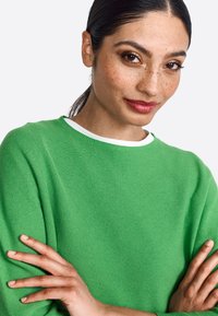 Groene gebreide trui met een textuurafwerking en een witte kraag. Het model heeft de armen gekruist, wat de pasvorm en stijlkenmerken van de trui laat zien.