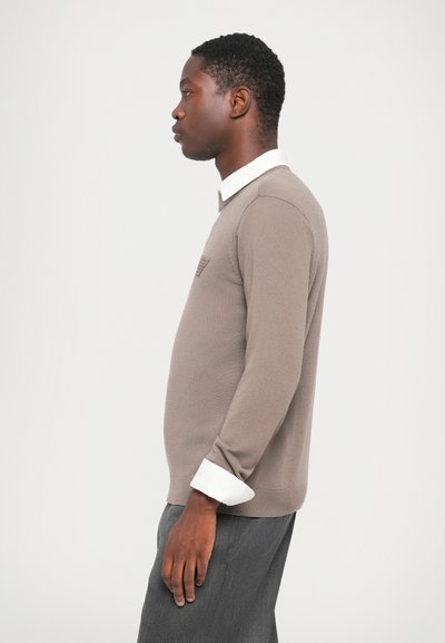 Brown knit sweater with a subtle pattern on the chest. Features a white collared shirt underneath and ribbed cuffs. Worn with gray trousers.