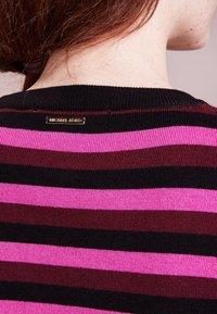 Maglione a righe con bande orizzontali in nero, rosso scuro e rosa. Presenta una texture a coste e una placca con logo dorato sul colletto.
