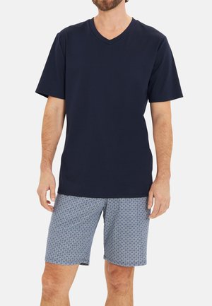 Mann trägt ein marineblaues V-Ausschnitt-Kurzarmshirt und blaue gemusterte Shorts und steht vor einem schlichten Hintergrund.