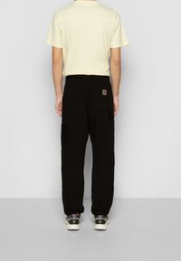 Svarta cargo byxor med flera fickor, parat med en ljusgul t-shirt. Byxorna har en avslappnad passform och subtil branding på baksidan.