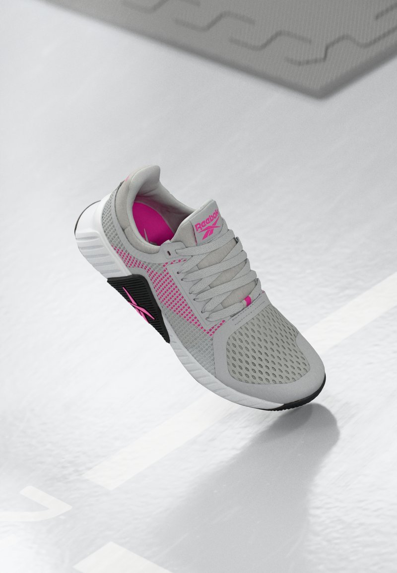 Pelēki un rozā Reebok sporta kedas ar sieta augšdaļu un detalizētu zoli, novietotas uz atstarojošas baltas virsmas.