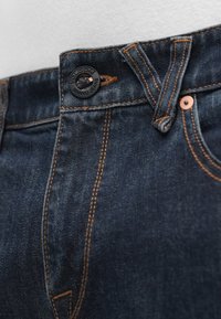 Μπλε τζιν από τζιν ύφασμα με χάλκινα ραφές, διαθέτει μαύρο κουμπί Volcom και φερμουάρ. Το ύφασμα φαίνεται υφασμένο και ανθεκτικό.