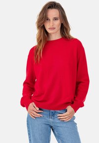 Pull en maille rouge avec un col rond et des manches longues ; texture douce ; porté par-dessus un jean en denim bleu clair.