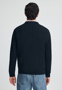 Zwart kabelgebreide trui met geribbelde manchetten en zoom, met een textuurpatroon. Draagt over lichtblauwe jeans, tegen een effen achtergrond.