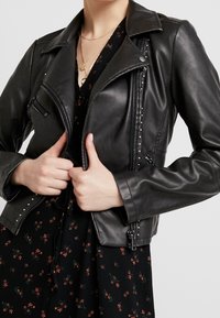 Giubbotto da motociclista in pelle nera con tasche con zip, dettagli borchiati e colletto a punte, indossato sopra un vestito floreale scuro.