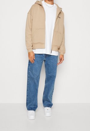 Beige zip-up hoodie met een voorzak, gedragen over een wit T-shirt, gecombineerd met blauwe spijkerbroek en witte sneakers.