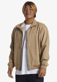 Tan lightweight jacket with a high collar, front zipper, and elastic cuffs. Worn over a white shirt, features a subtle logo detail.