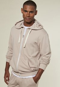 Light beige zip-up hoodie with drawstrings, kangaroo pocket, ribbed cuffs, and hem. Worn over a white t-shirt with a soft texture.