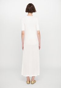 Robe longue blanche en tricot côtelé avec des manches courtes, présentant une texture lisse et une jupe fluide. Des chaussures dorées à enfiler sont portées.